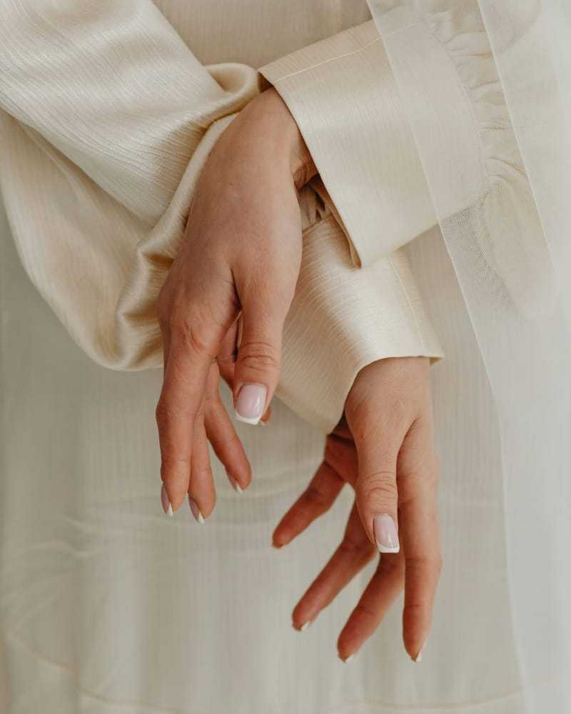 Hands with neatly manicured nails, wearing a cream-colored, textured fabric garment.