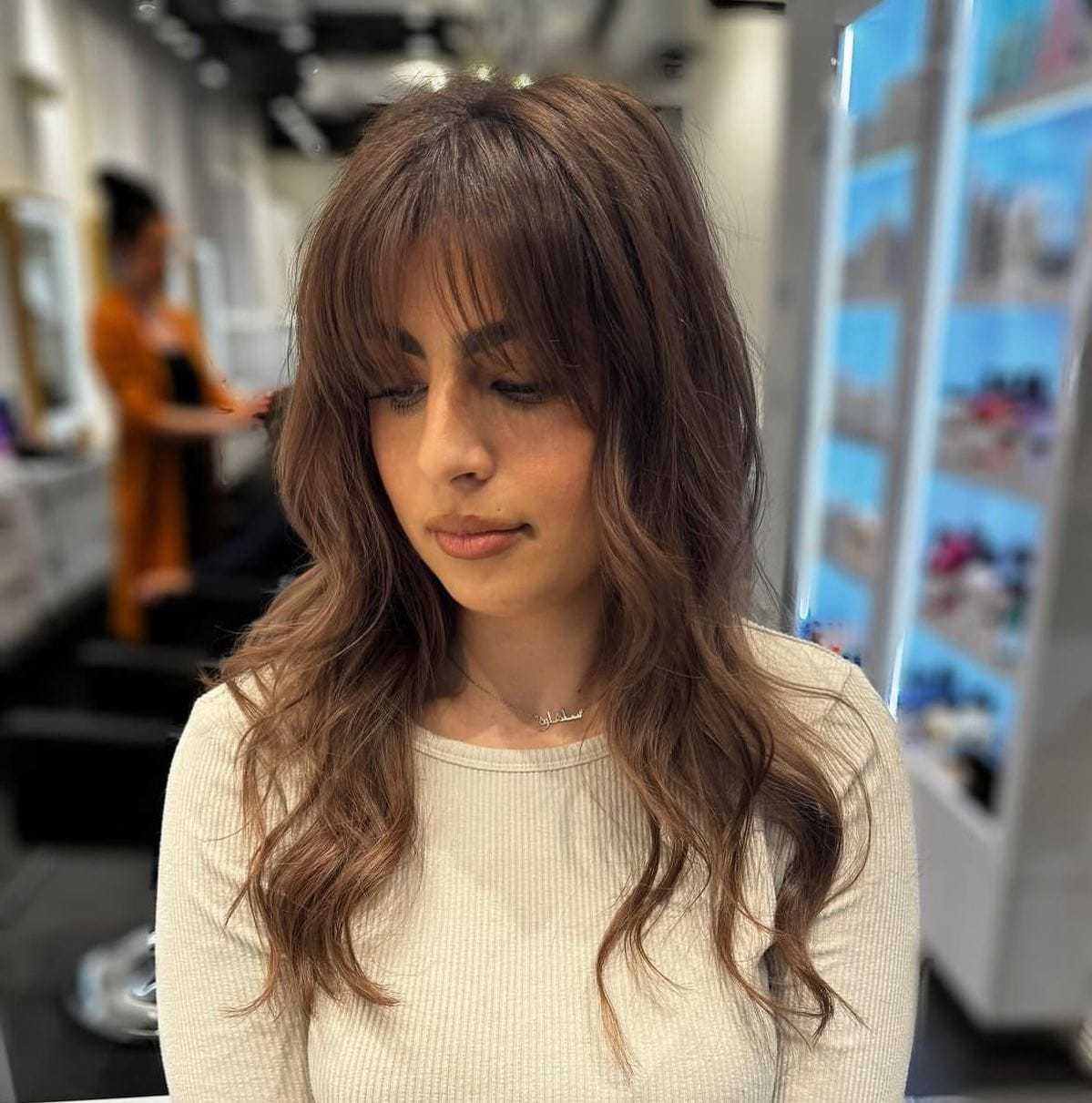 Woman with wavy brown hair and bangs, wearing a white top in a salon setting.