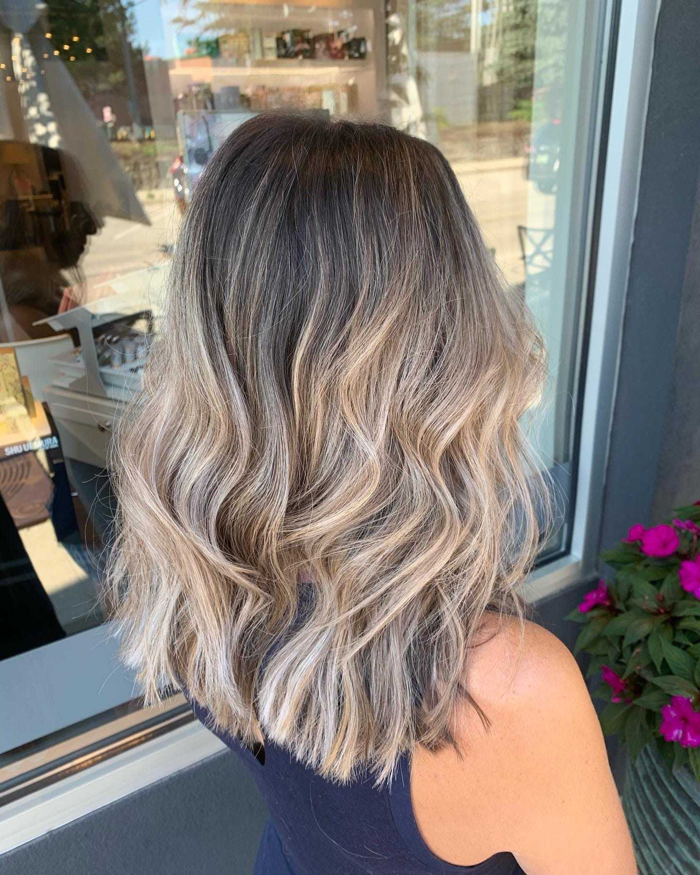 Wavy blonde balayage hairstyle on woman, viewed from behind, standing outside a salon.
